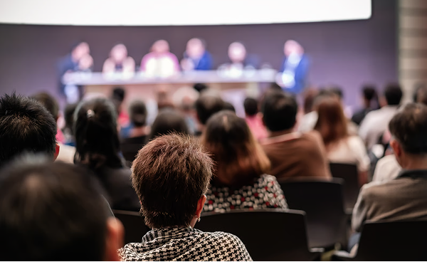 A panel of speakers seated at the front of a conference room, seen from behind the audience